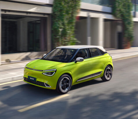 Dzeltens pilsētas elektroauto Dongfeng Box ar baltu jumtu brauc pa pilsētu, skats no priekšas un sāniem
